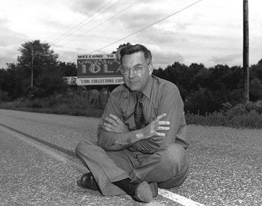 Честер Краузе сидит на дороге у таблички своего города Айола, где была основана его фирма.